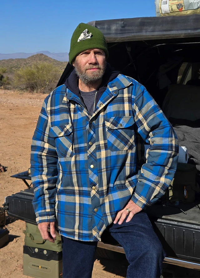 Front view of a man in a blue khaki men's quilted plaid flannel hoodie leaning against the rear of a car