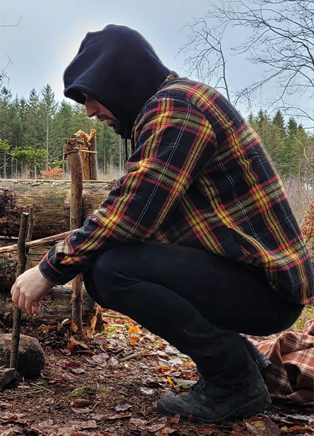 Side view of a man wearing a red and black men's sherpa lined jacket crouching on the ground with a hat on
