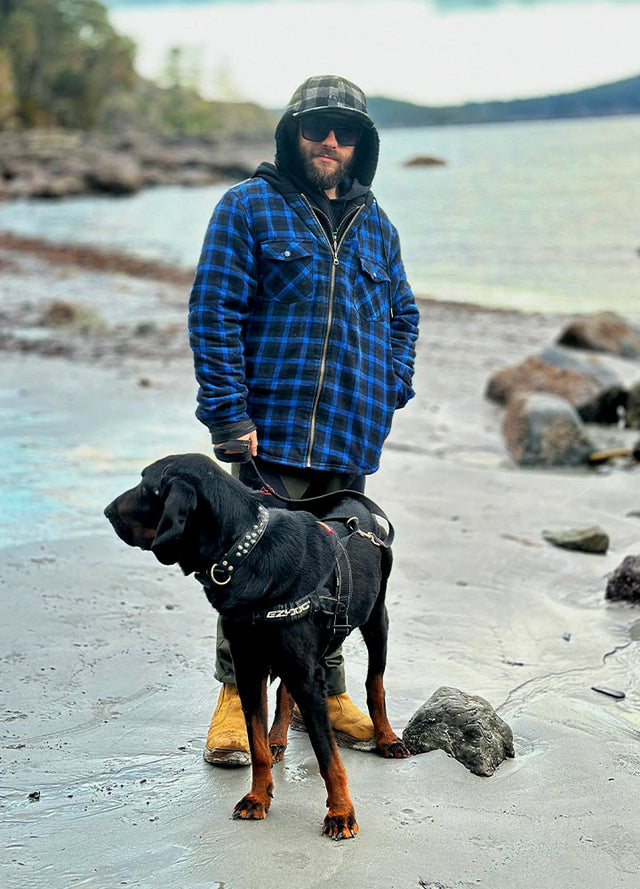 Front view of a man wearing a dark blue men's hooded flannel plaid jacket and leading his dog by the sea