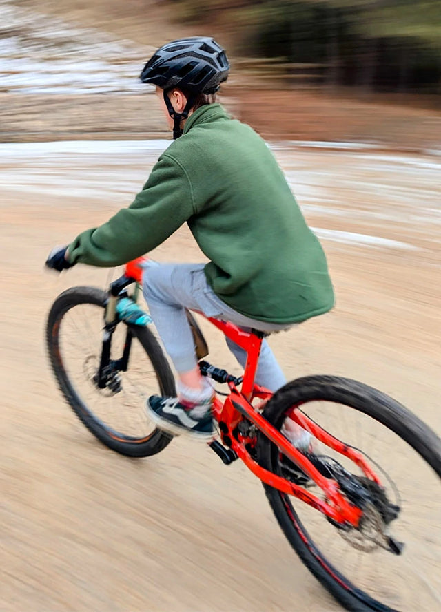 A boy in an army green plaid fleece shirt jacket rides his mountain bike along a muddy path.