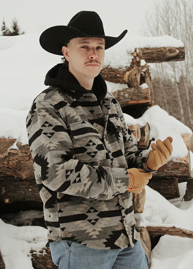 Man in a patterned Aztec jacket and cap outdoors, holding a knife.