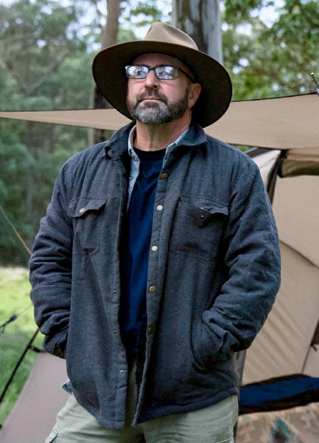 Front view of a man in a dark gray men's sherpa lined plaid shirt jacket standing in front of the tent