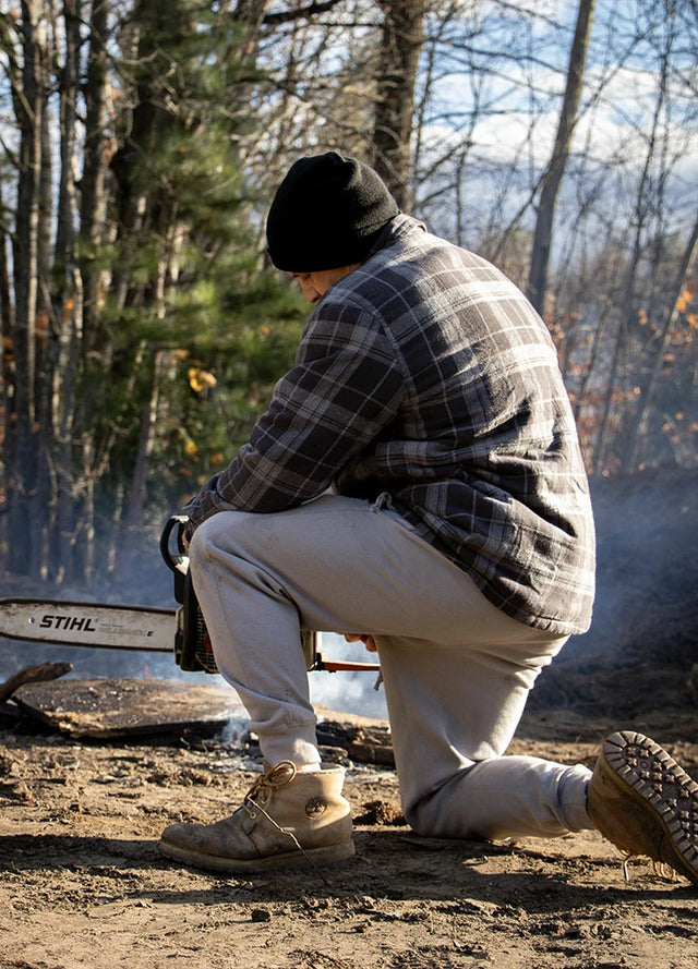 Back view of a man wearing a gray men's warm flannel shirt jacket crouching on the ground with a chainsaw