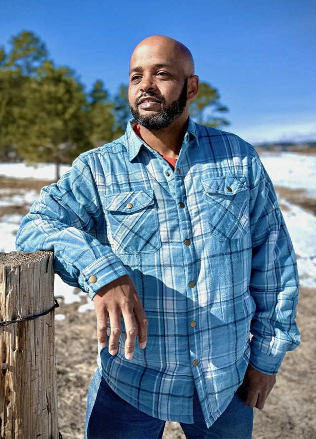 Front view of a man in a light blue men's sherpa lined flannel plaid jacket leaning on a wooden stake