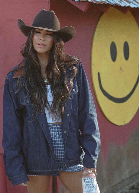 Front view of woman in a dark blue denim flannel men's shirt and jacket standing in front of a wall