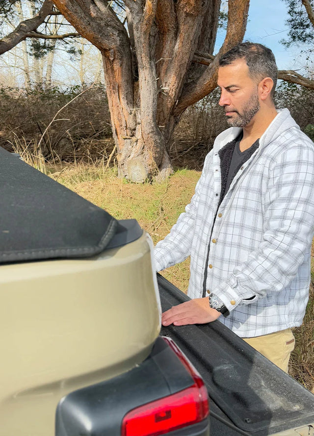 Side view of a man wearing a white and gray men's flannel hooded jacket standing at the rear of a car