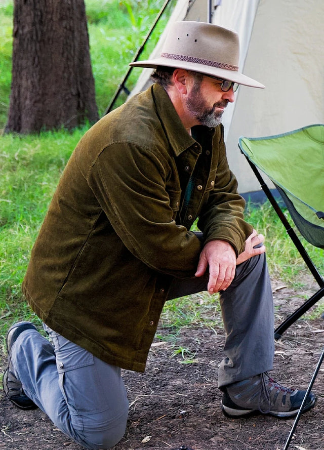 Side view of a man wearing a men's army green flannel-lined corduroy shirt jacket, kneeling outdoors while waiting for his coffee to brew during a camping trip