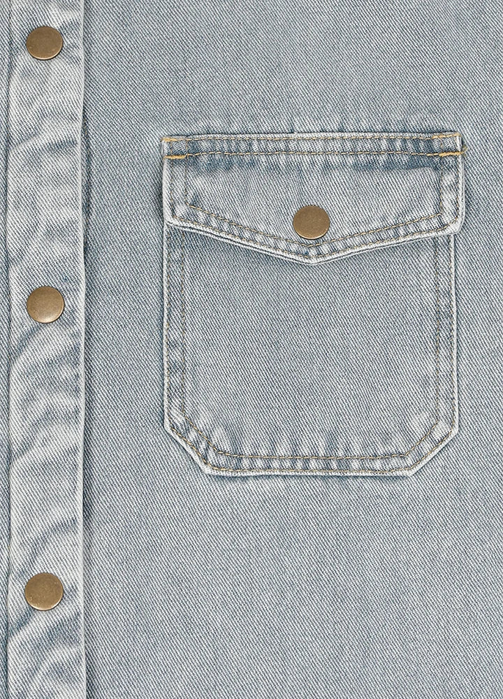 Close-up of a light blue denim button-up shirt featuring a chest pocket with a flap and a brass snap button.
