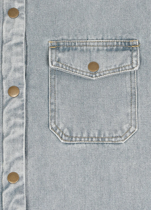 Close-up of a light blue denim button-up shirt featuring a chest pocket with a flap and a brass snap button.
