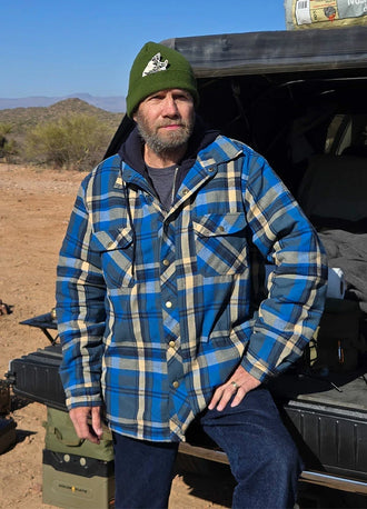 Front view of a man in a blue khaki men's quilted plaid flannel hoodie leaning against the rear of a car