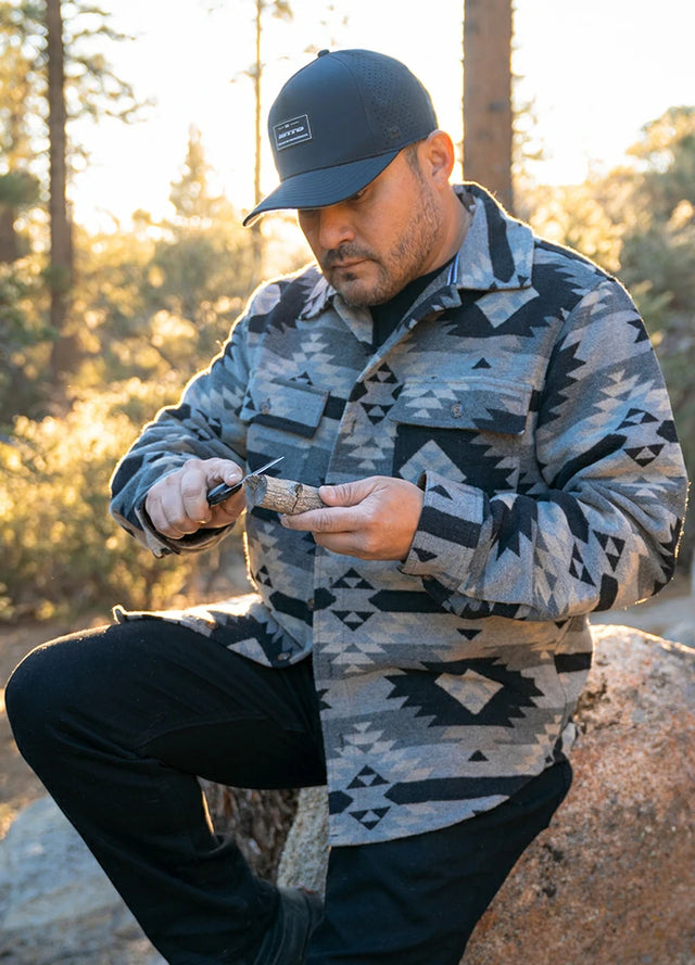 Man in a patterned Aztec jacket and cap outdoors, holding a knife.