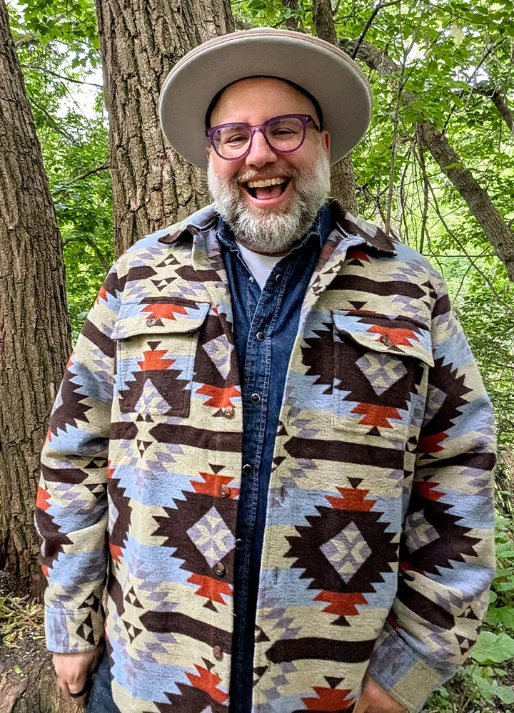 Front view of a man wearing a Coffee and Blue men's Aztec shirt jacket, hands in pockets, smiling warmly and laughing