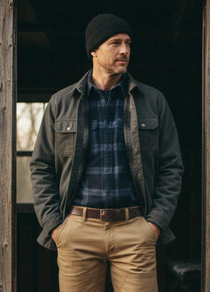 A man wearing a dark charcoal overshirt, blue and gray plaid flannel shirt, tan pants, and a black beanie, standing in a rustic wooden doorway-XR123GS0S,XR123GS0M,XR123GS0L,XR123GS0XL,XR123GS0XXL