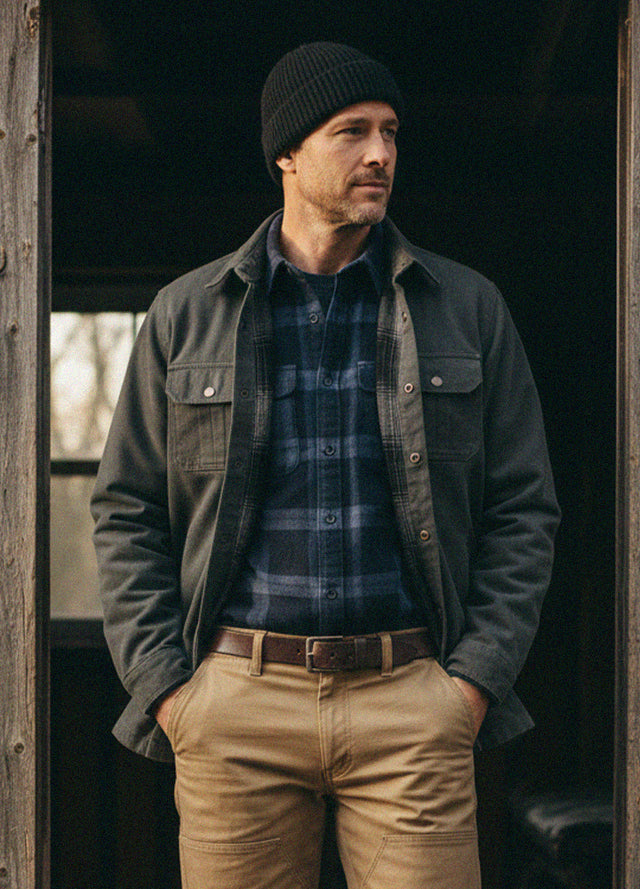 A man wearing a dark charcoal overshirt, blue and gray plaid flannel shirt, tan pants, and a black beanie, standing in a rustic wooden doorway-XR123GS0S,XR123GS0M,XR123GS0L,XR123GS0XL,XR123GS0XXL