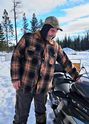Front view of a man in a brown black men's quilted lined brushed flannel hooded jacket standing next to the sled