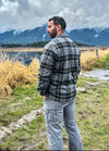 Back view of a man wearing a black and gray men's fleece lined plaid shirt jacket standing in the grass