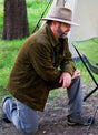 Side view of a man wearing a men's army green flannel-lined corduroy shirt jacket, kneeling outdoors while waiting for his coffee to brew during a camping trip