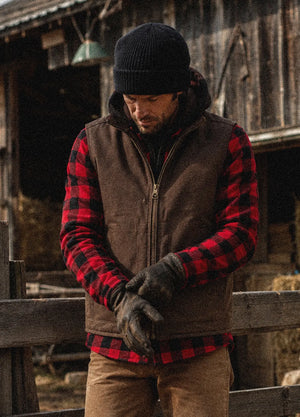 A man wearing a brown zip-up vest over a red and black buffalo plaid flannel shirt, a black knit beanie, and dark gloves, standing in front of a rustic wooden barn.